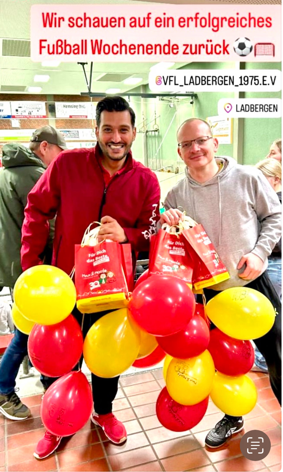 Zwei Männer lächelnd mit roten Jacken und roten sowie gelben Luftballons in einem Geschäft.