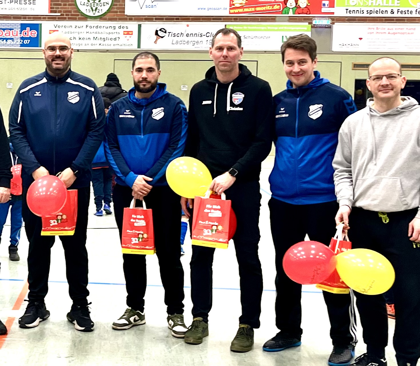 Fünf Männer lächeln, halten rote Geschenktüten und Luftballons in einer Sporthalle.